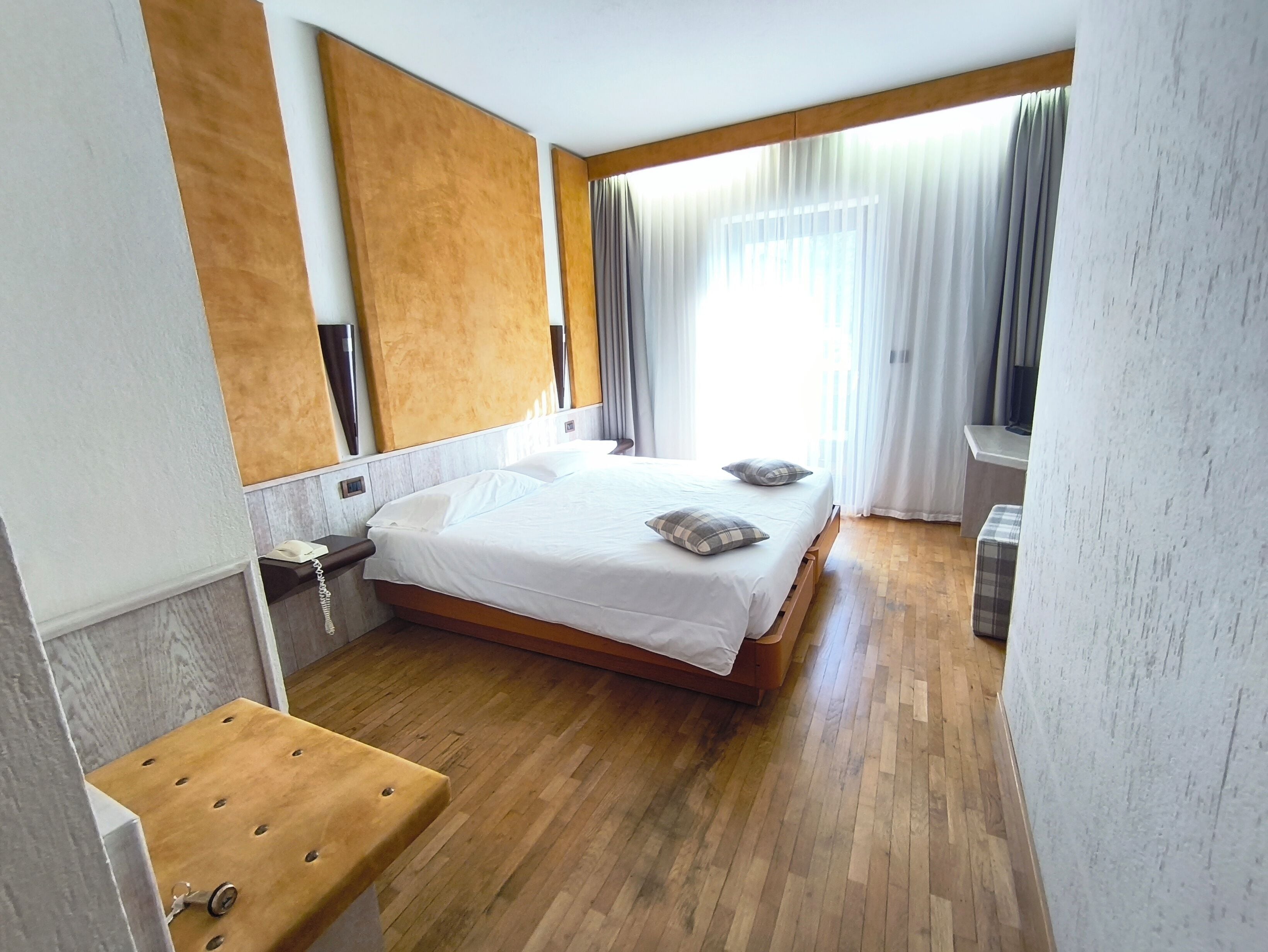 A hotel room with a wooden bed, a wooden bench, a small table, a mirror, and a window with curtains.