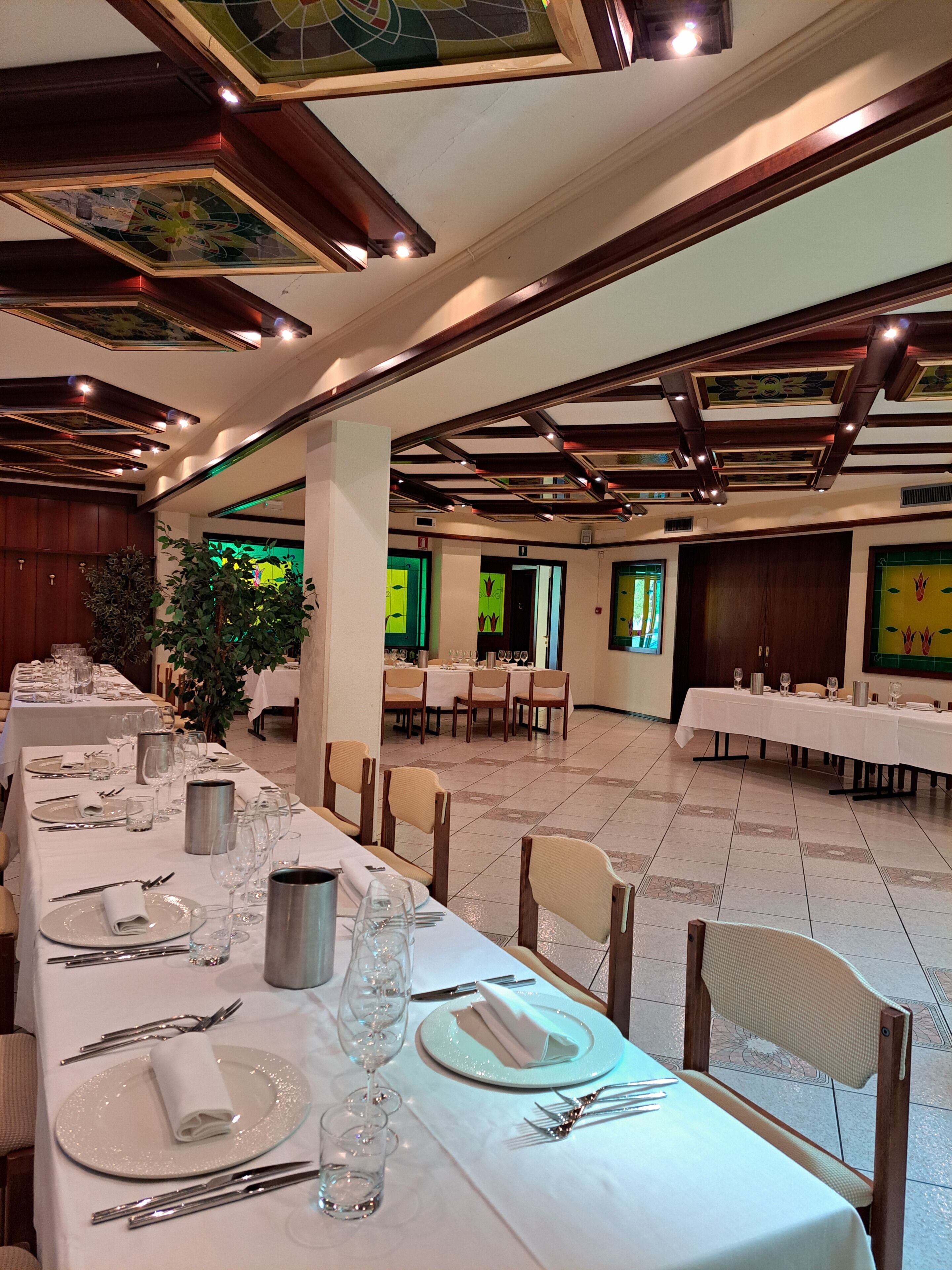 A banquet hall with long tables set for dining, wooden beams on the ceiling, and a large plant in the center.