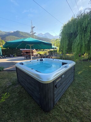Un jacuzzi en un área verde con montañas al fondo, mobiliario exterior y un edificio con toldo verde.