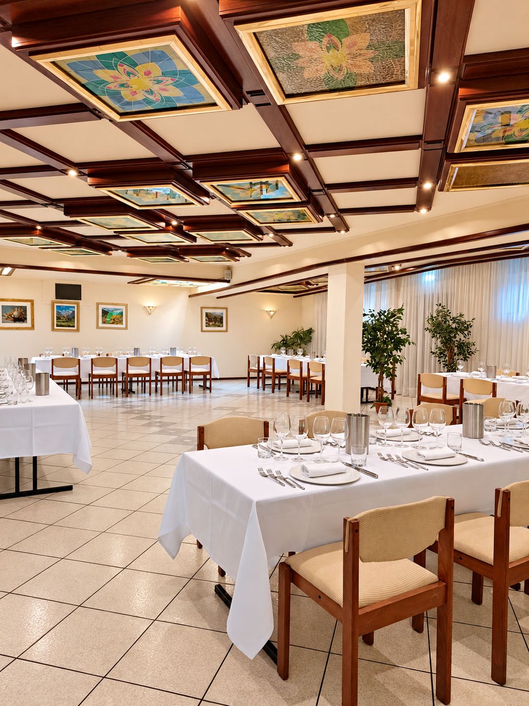A spacious dining area with multiple tables set for a meal, featuring a high ceiling with decorative panels and a tiled floor.