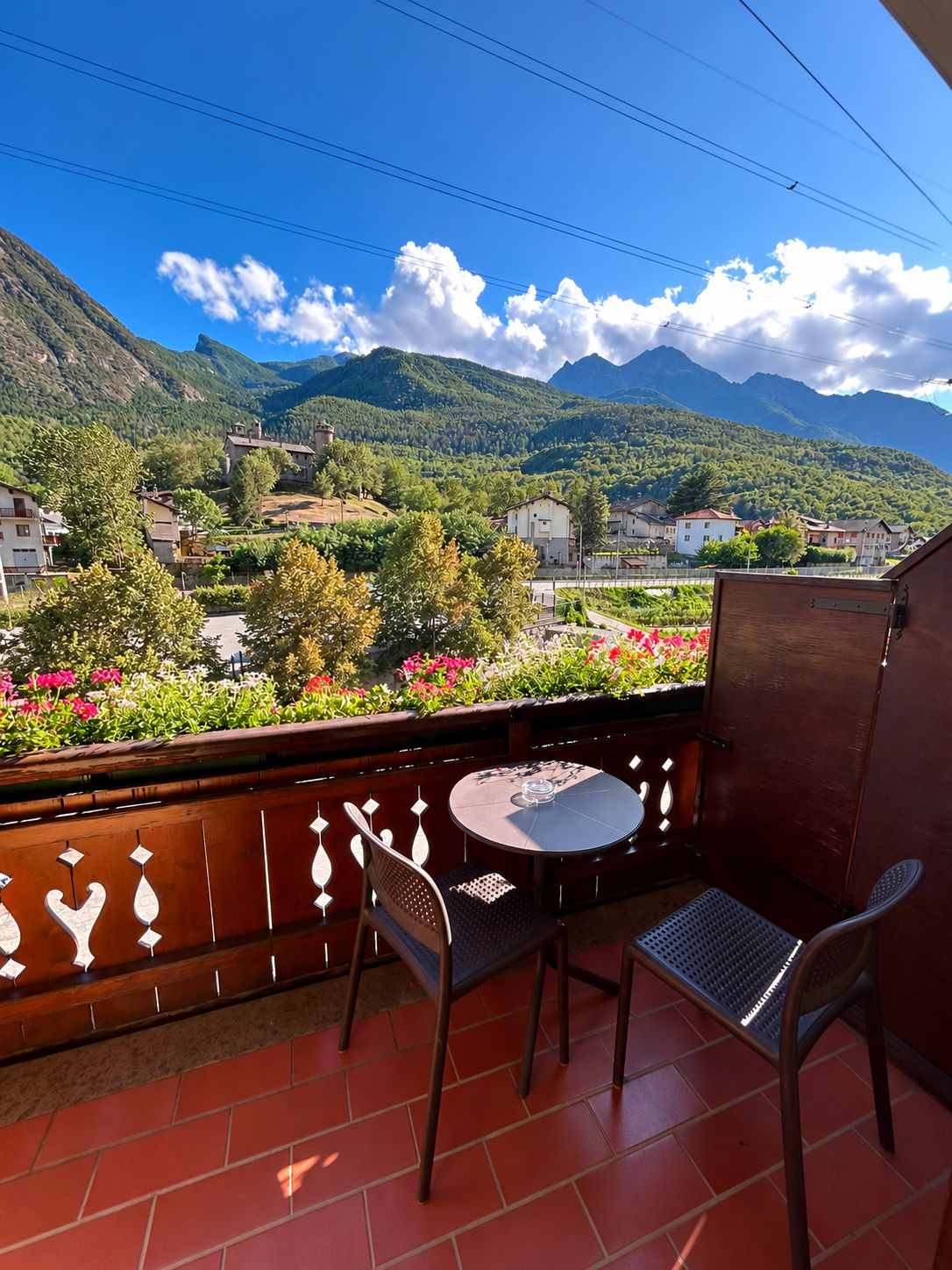 Un balcón con una mesa redonda y sillas, con vistas a un paisaje montañoso con casas y vegetación.
