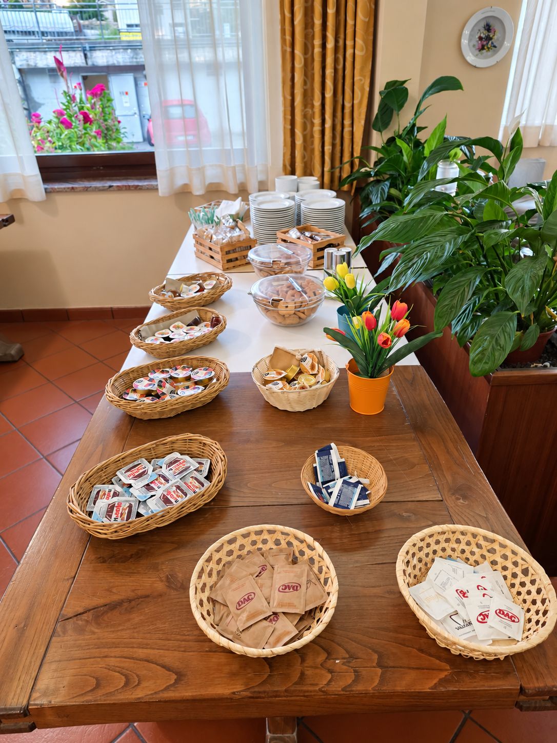 A buffet table with various food items and baskets, a potted plant with tulips, and a window with curtains.
