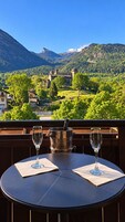 Una mesa con dos copas de vino en un balcón con vista a un paisaje montañoso que incluye un castillo.