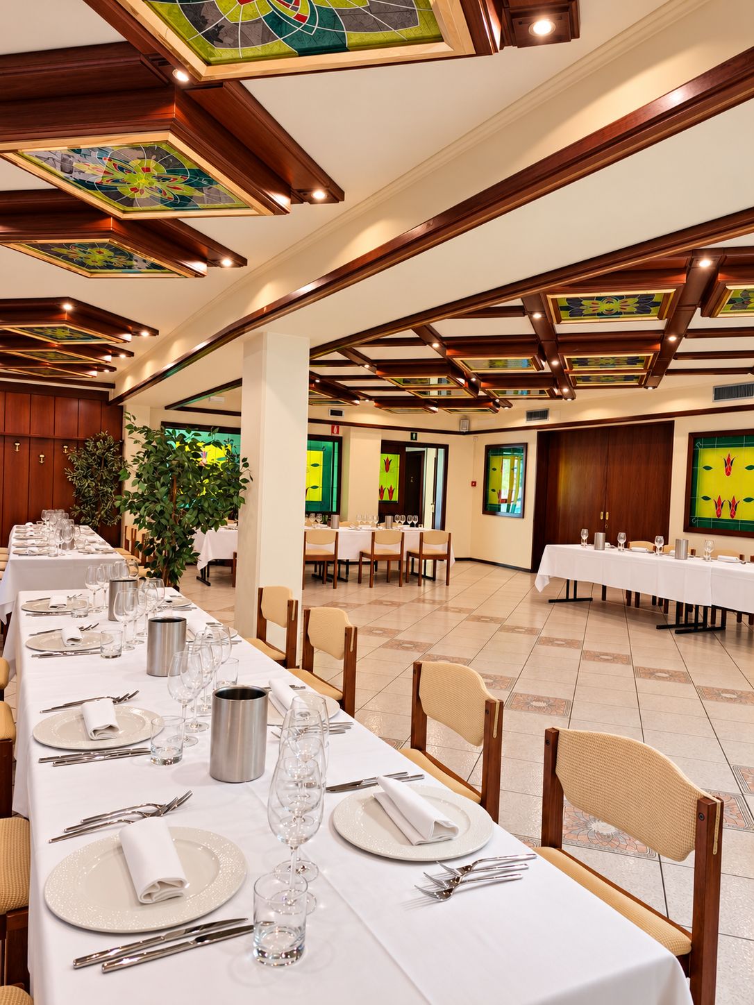 Een feestzaal met lange tafels klaargeset voor een diner, houten balken in het plafond en een grote plant in de hoek.