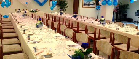 Un salón de banquetes con largas mesas dispuestas para un evento formal, decorado con globos azules y blancos y centros de mesa florales.