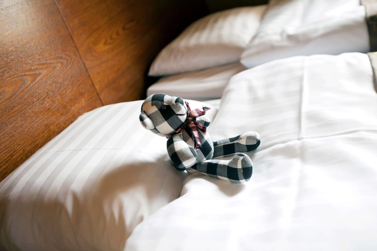 Una cama con sábanas blancas y un oso de peluche a cuadros sobre ella.