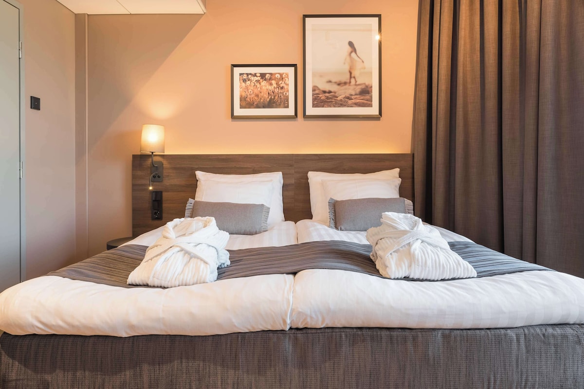 Una cama bien hecha con ropa de cama blanca y gris, dos cuadros en la pared y una lámpara en la mesita de noche.