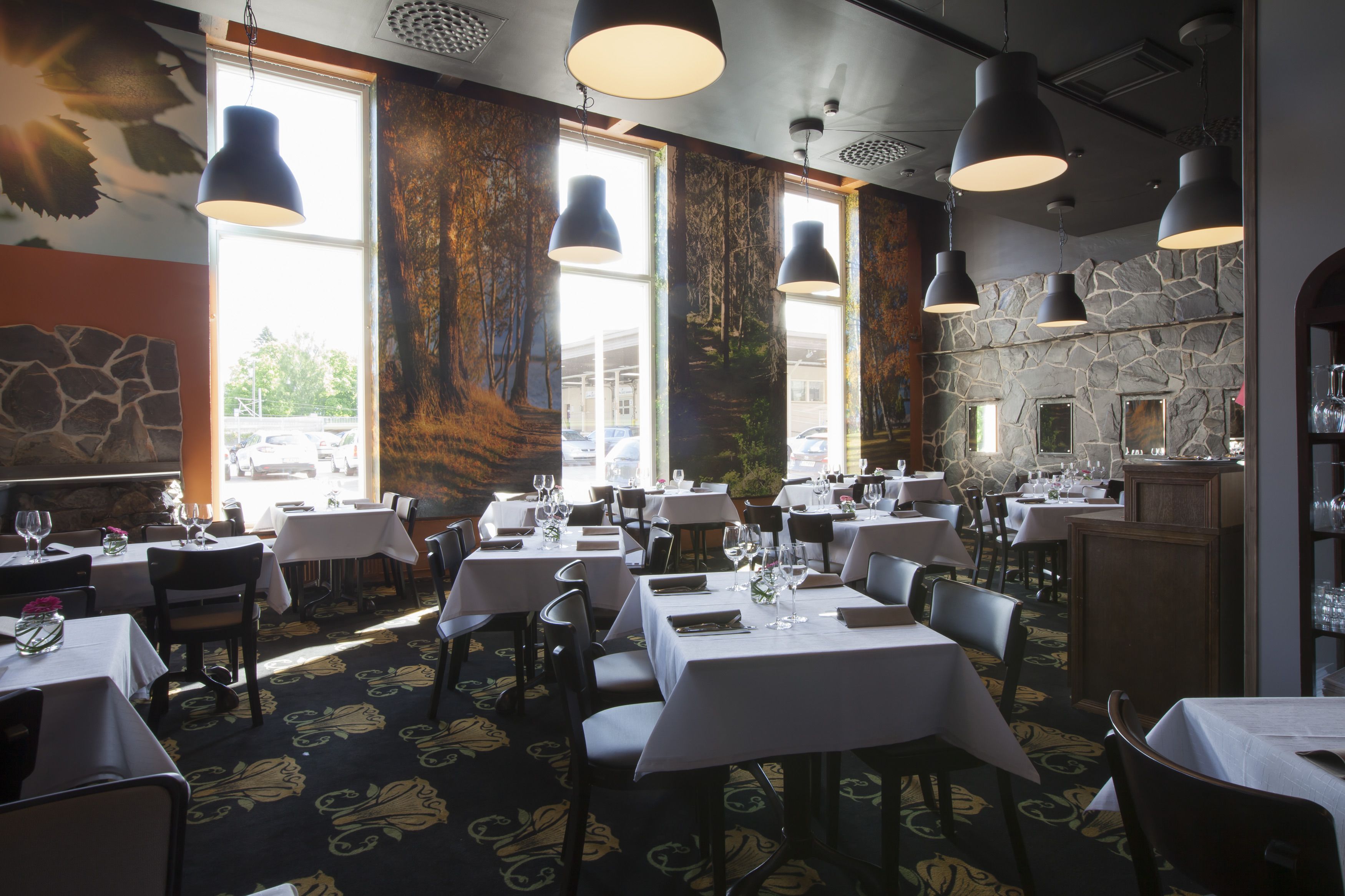 A restaurant interior with multiple tables, hanging pendant lights, and a stone wall.