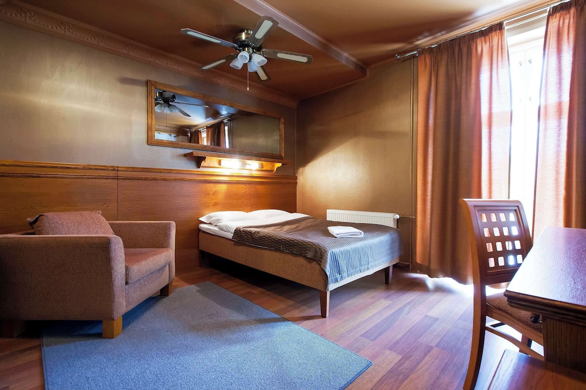 Una habitación de hotel con cama, una silla, un escritorio, ventilador de techo, espejo y ventana con cortinas.