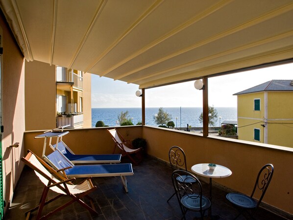 Un balcón con vistas al mar, sillones reclinables azules, una mesa con sillas y una mesita redonda con un cuenco.