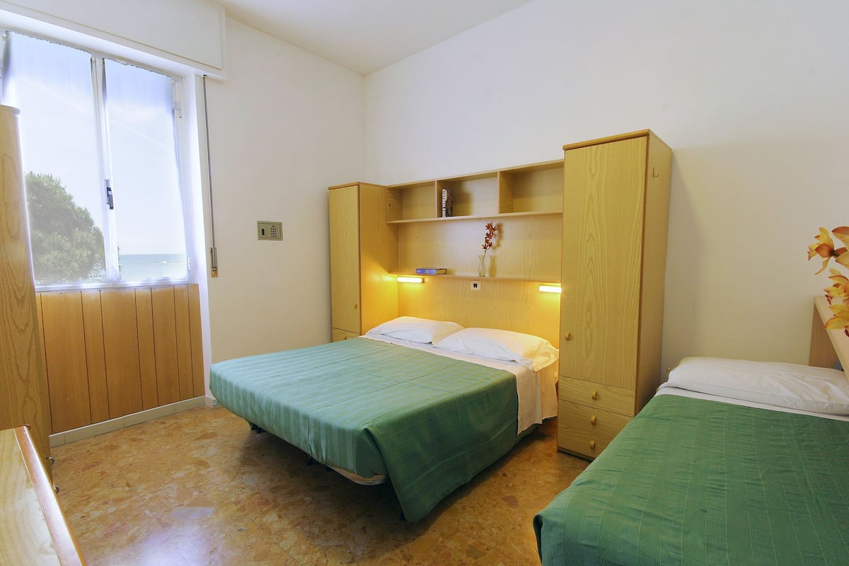 Ein Zimmer mit zwei Betten, einem Holzschrank, einem kleinen Tisch und einem Fenster mit Meerblick.