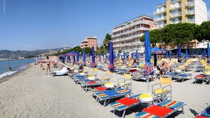 A beach with rows of colorful beach chairs and umbrellas, a clear sky, and buildings in the background. - Hotel Anita (San Bartolomeo al Mare)