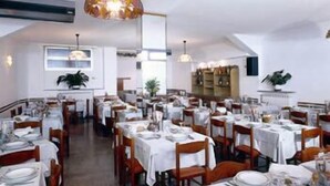 A dining area with multiple tables set for service, a hanging light fixture, and a kitchen area in the background. - Hotel Anita (San Bartolomeo al Mare)