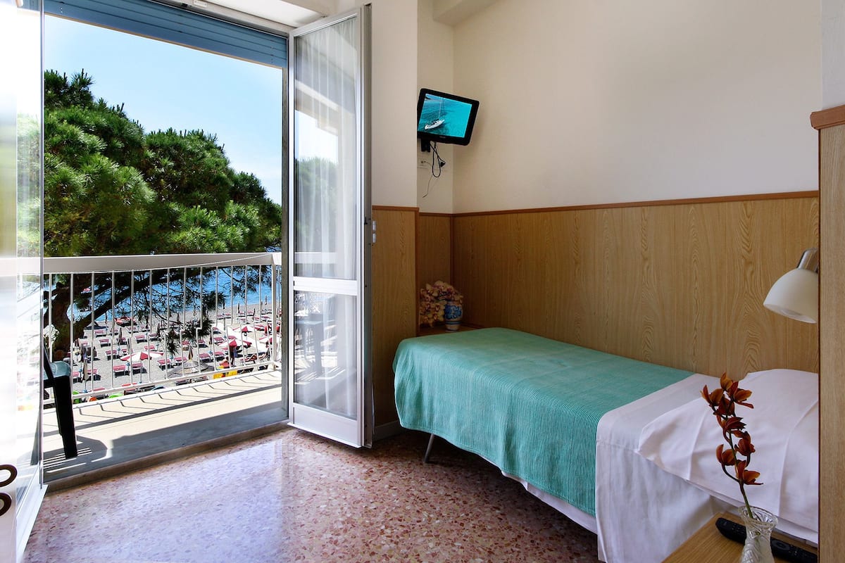 Ein Hotelzimmer mit Bett, Wandfernseher und einem Balkon mit Blick auf Strand und Palmen.