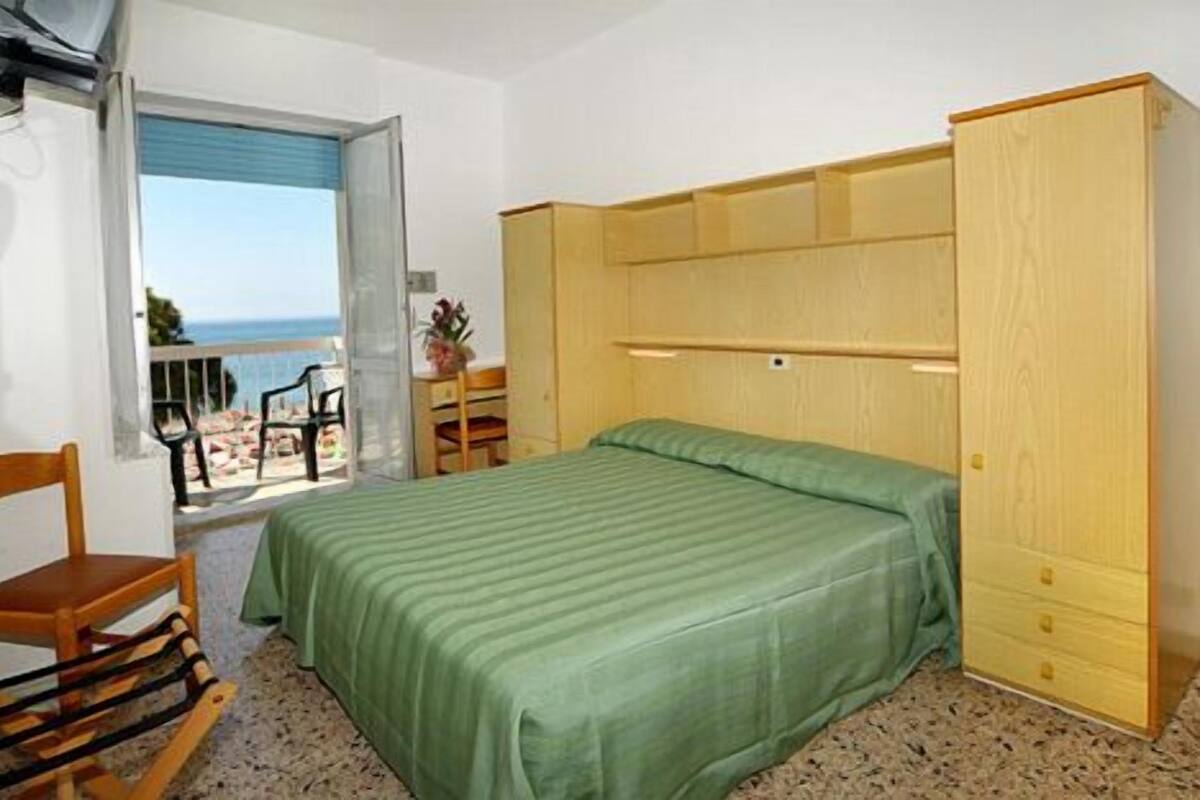 Ein Schlafzimmer mit einem Bett, einem Holzschrank, einem Schreibtisch und einem Stuhl. Ein Balkon bietet Blick auf das Meer.