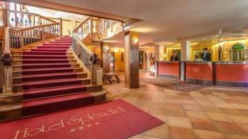 Un'ampia hall d'albergo con una maestosa scala, una reception e un tappeto rosso con il nome dell'hotel.