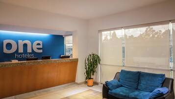 Ein Hotel-Empfangsbereich mit einer Rezeption, einem blauen Sofa, einer Zimmerpflanze und einem großen Fenster.