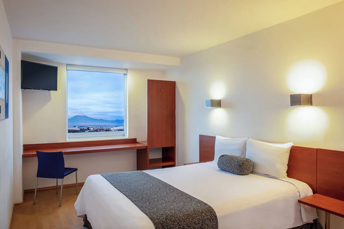 A hotel room with a bed, a desk, a chair, a window with a view, and wall-mounted lights.