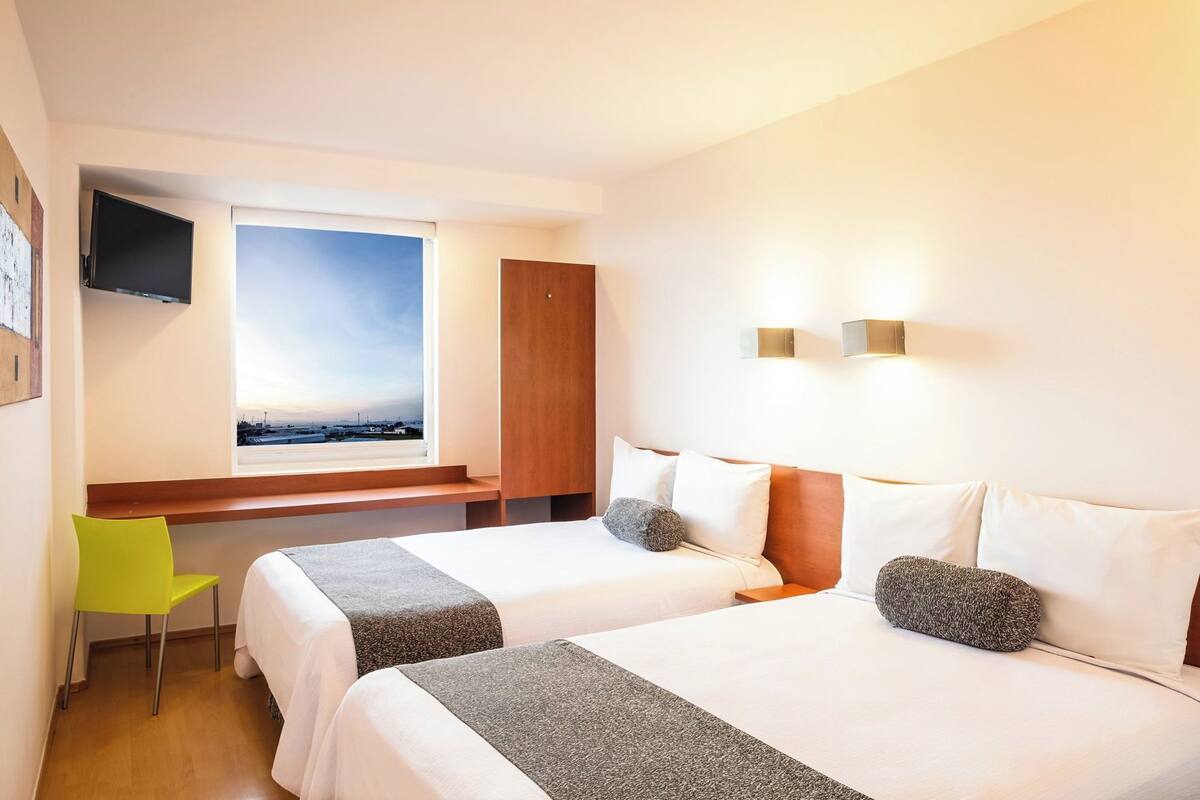 A hotel room with two beds, a desk, a chair, a TV, and a window with a view.