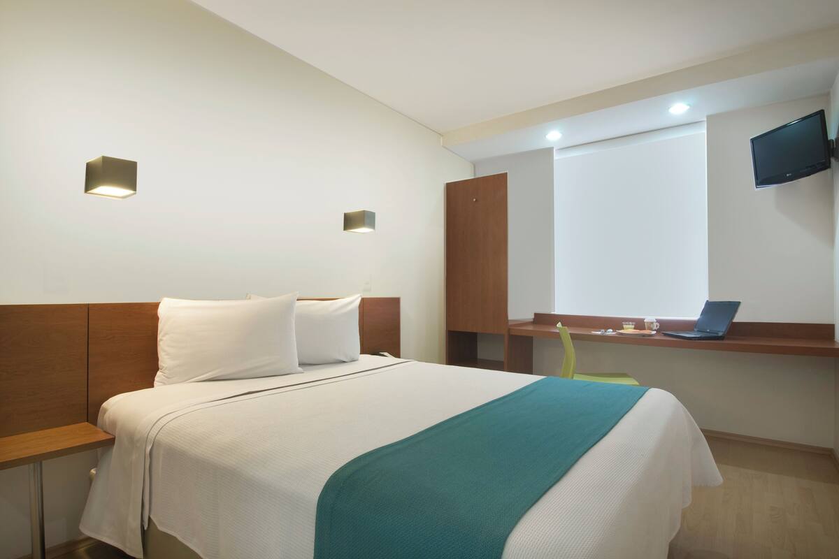 A hotel room with a bed, a desk with a laptop, a TV mounted on the wall, and a small shelf with a cup and a book.