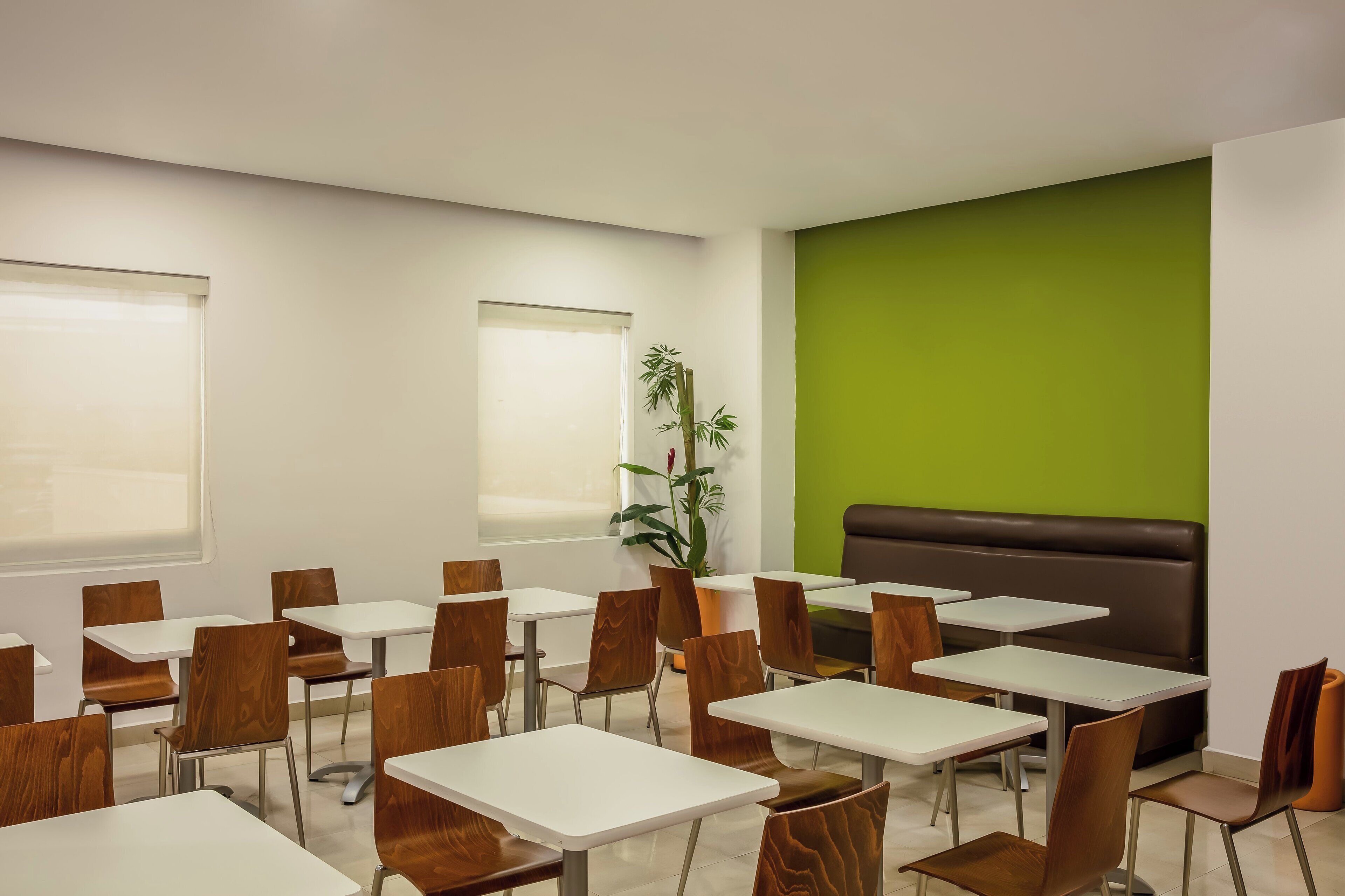 A modern dining area with white tables and wooden chairs, a green accent wall, and a large window.