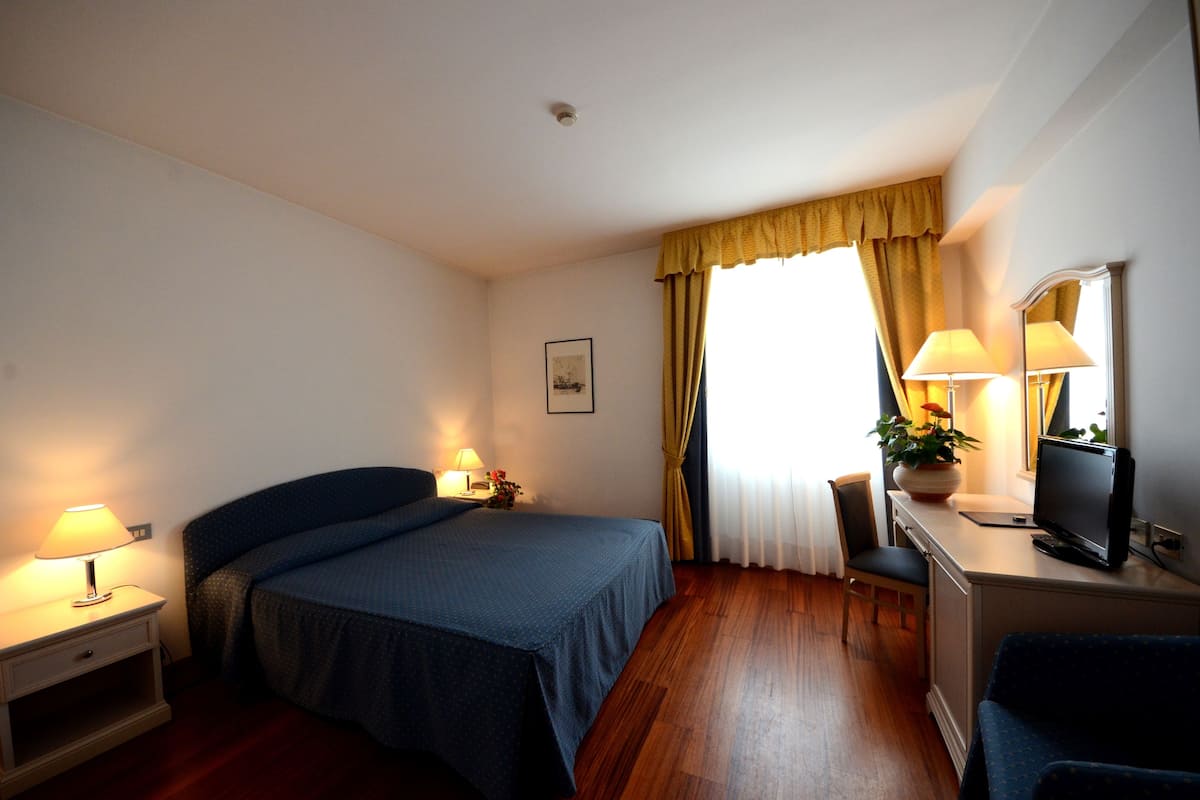Habitación de hotel con cama, lámparas de noche, escritorio con computadora, silla y ventana con cortinas.