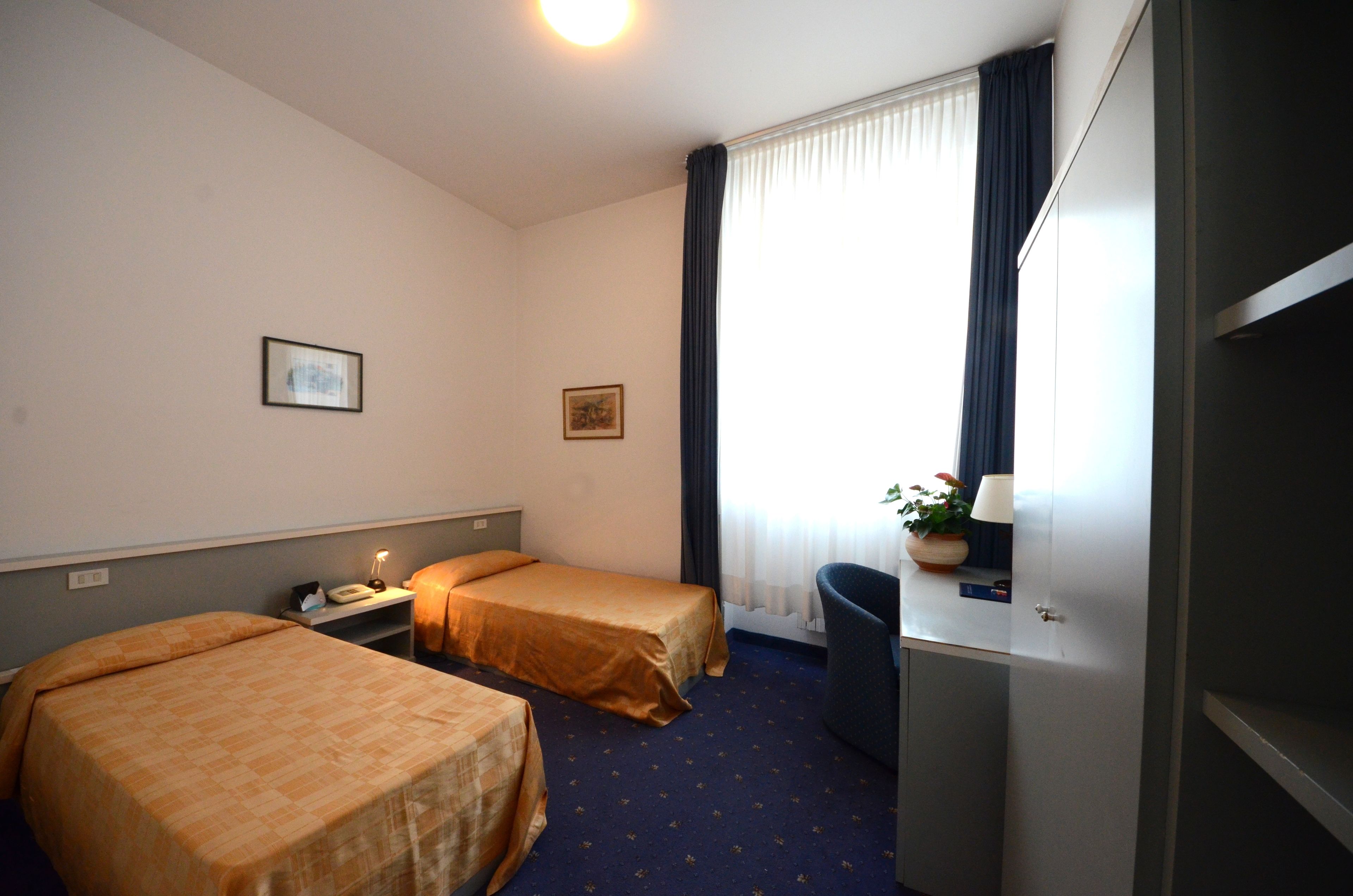 Una camera d'albergo con due letti, una scrivania, una sedia, una lampada e una finestra con tende.