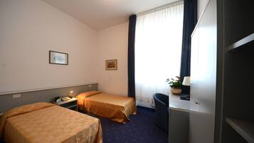 Una camera d'albergo con due letti, una scrivania, una sedia, una lampada e una finestra con tende.