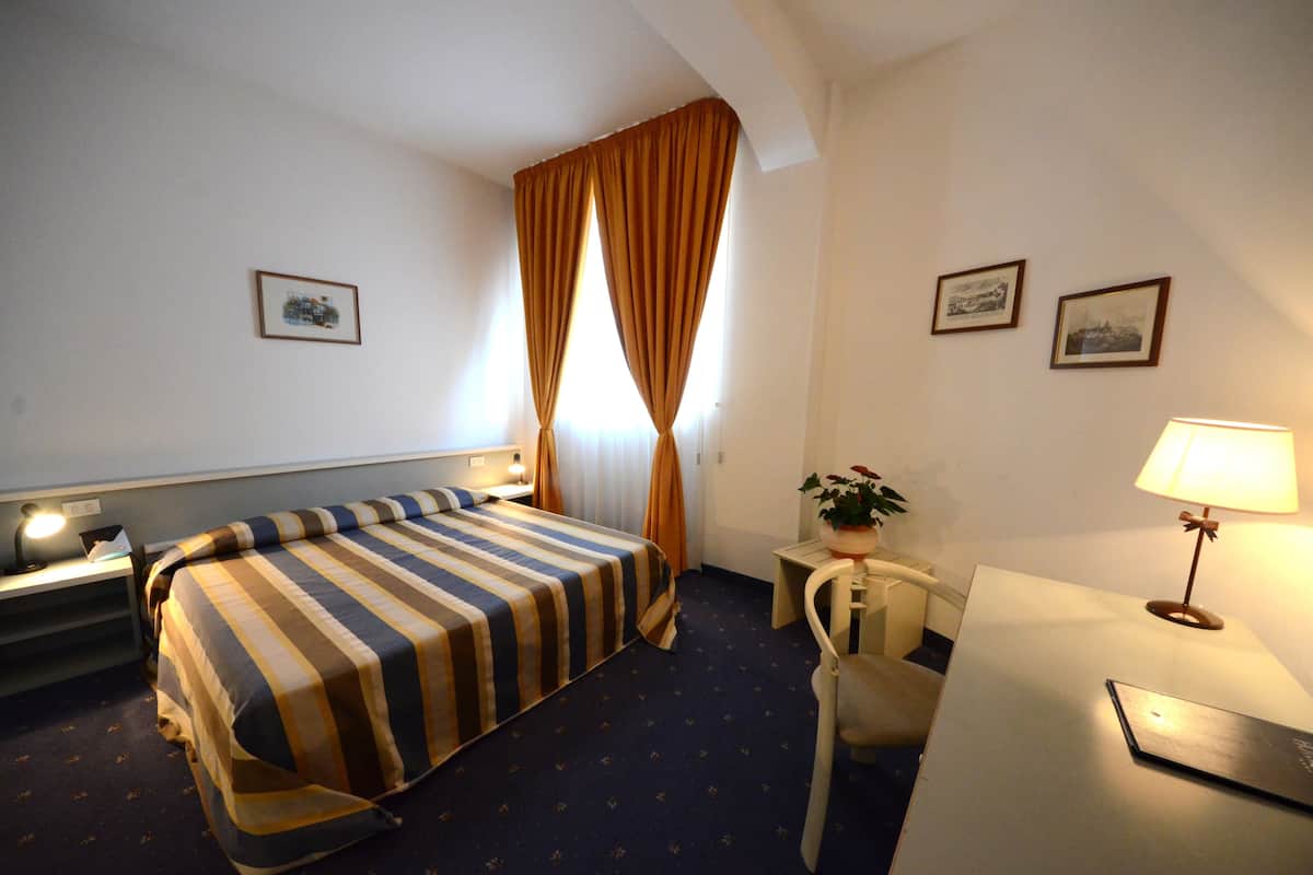 Habitación de hotel con cama, escritorio, silla, lámpara y dos cuadros en la pared.