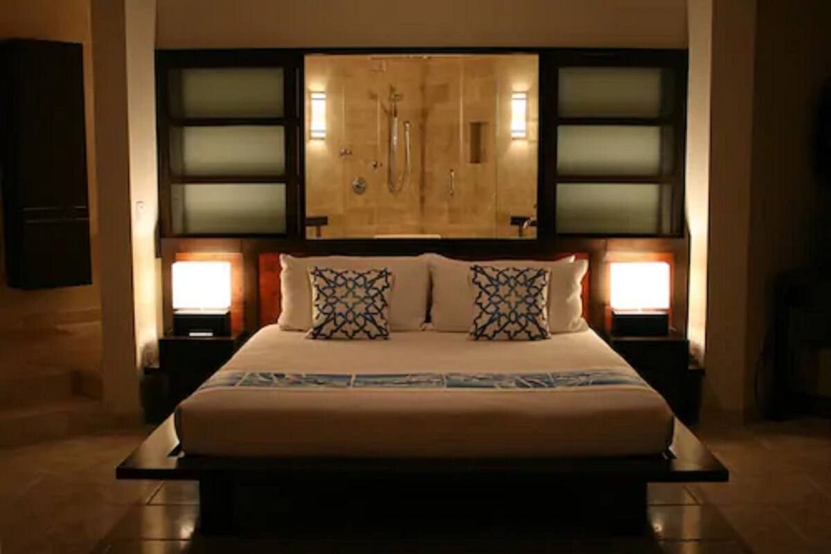 A large bed with patterned pillows, a headboard with a built-in shower, and two bedside lamps.