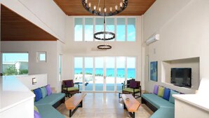 A spacious living area with a large window overlooking the beach, a ceiling fan, and a flat-screen TV mounted on the wall. - Sky Beach Club (Governor's Harbour)