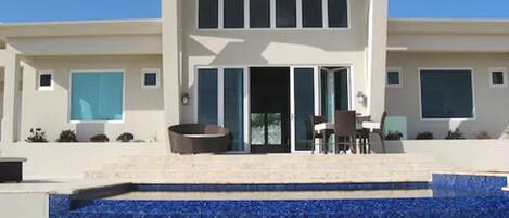 Una casa moderna de dos pisos con piscina, un patio con mobiliario exterior y un amplio ventanal con vista a la piscina.