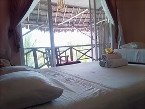 Habitación de hotel con una cama, almohadas, una pequeña mesa con una toalla enrollada y vistas a una estructura de techo de paja y follaje exterior.