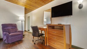 A room with a wooden ceiling, a purple recliner, a desk with drawers, a chair, a flat-screen TV mounted on the wall, and a floor lamp.