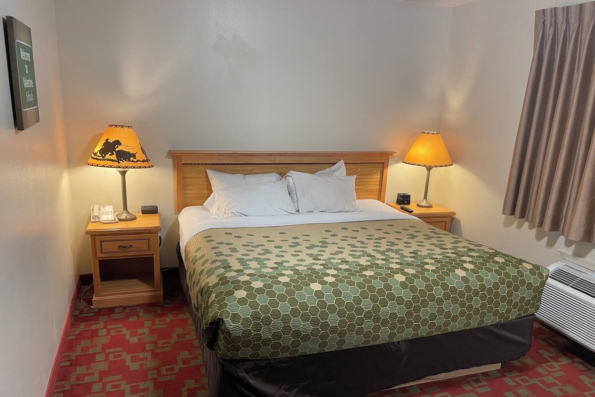A hotel room with a bed, two bedside tables, a telephone, a remote control, and a wall-mounted air conditioner.