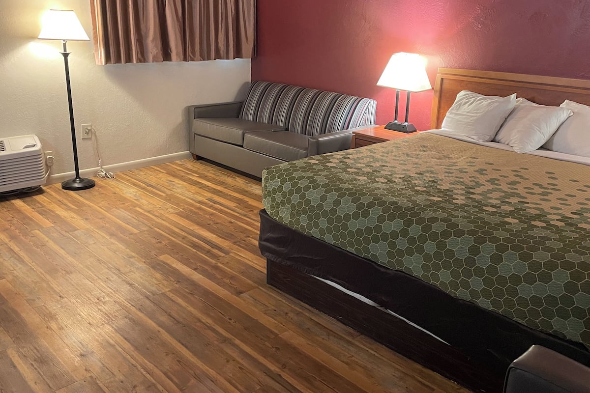 A hotel room with a bed, a sofa, a nightstand with a lamp, and a heater on the floor.