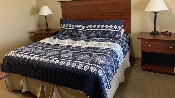 A neatly made bed with a patterned bedspread, two bedside tables with lamps, and framed artwork on the wall. - Corporate East Hotel (Ulysses)