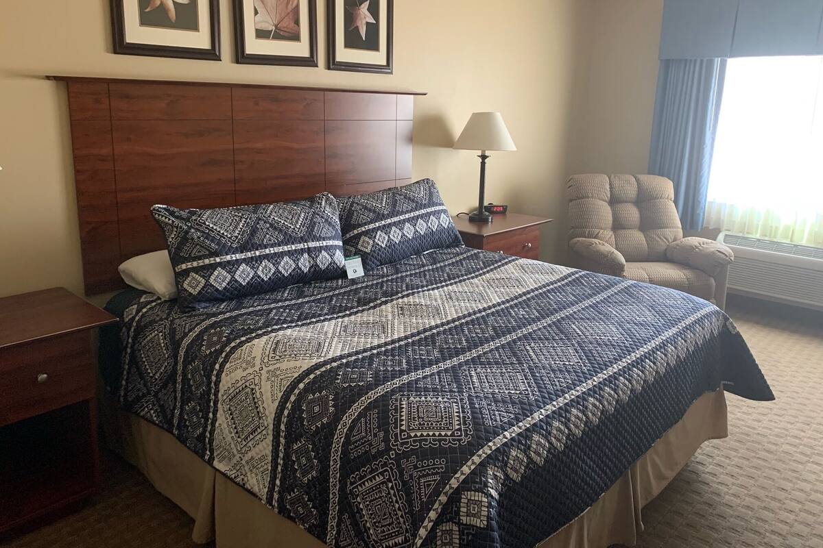 A hotel room with a large bed, a wooden headboard, a chair, a nightstand with a lamp, and framed pictures on the wall.