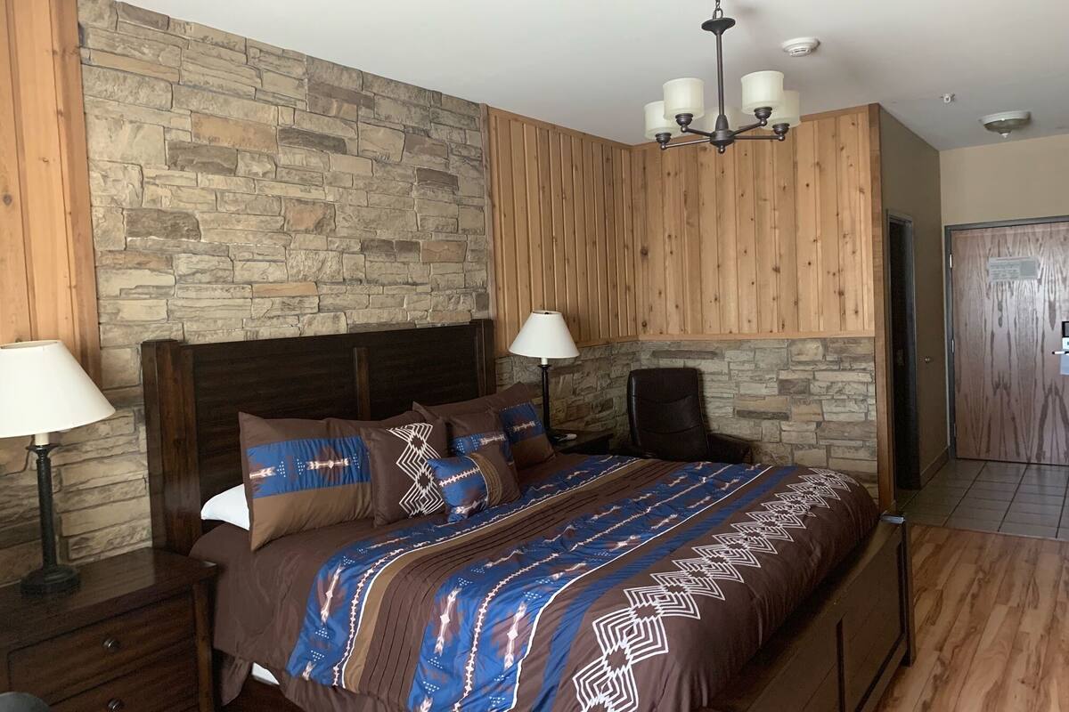 A bedroom with a stone wall, wooden paneling, a bed with patterned bedding, two bedside lamps, and a ceiling light fixture.