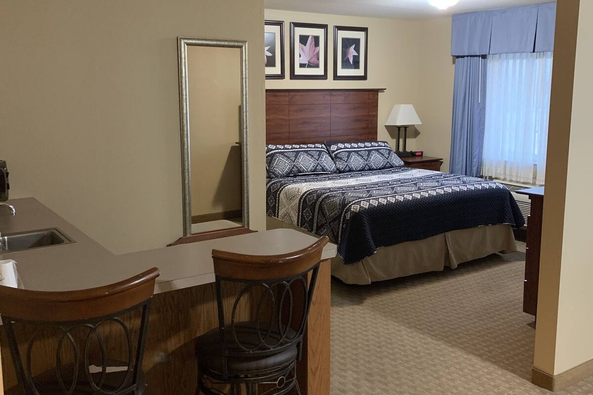 A hotel room with a bed, a desk with chairs, a mirror, a nightstand, a lamp, and a window with curtains.