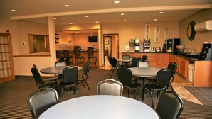 A hotel room with a dining area featuring round tables and chairs, a kitchen area with a coffee machine, and a television mounted on the wall.