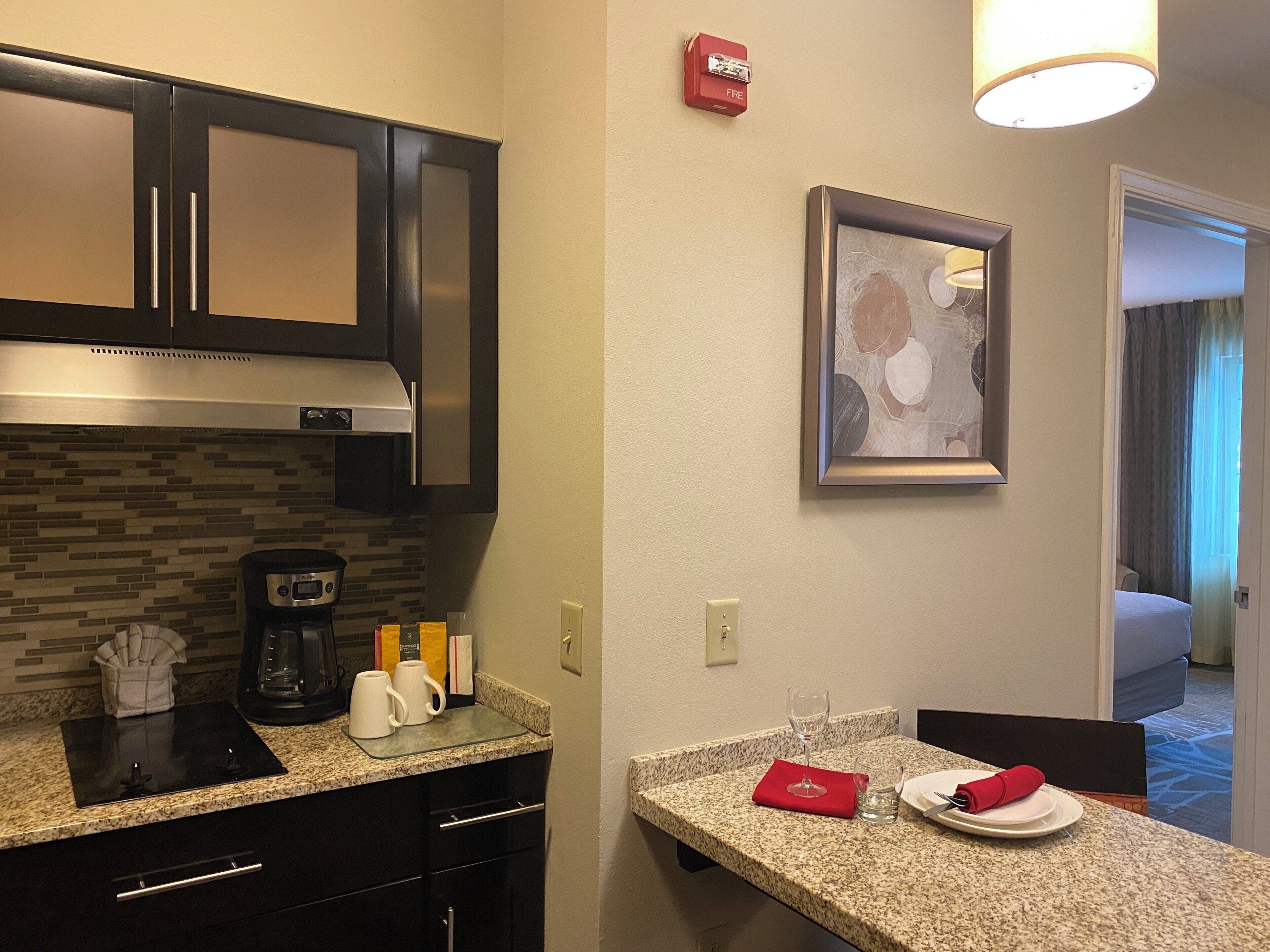 A hotel kitchen with a coffee maker, a framed artwork, and a fire alarm.