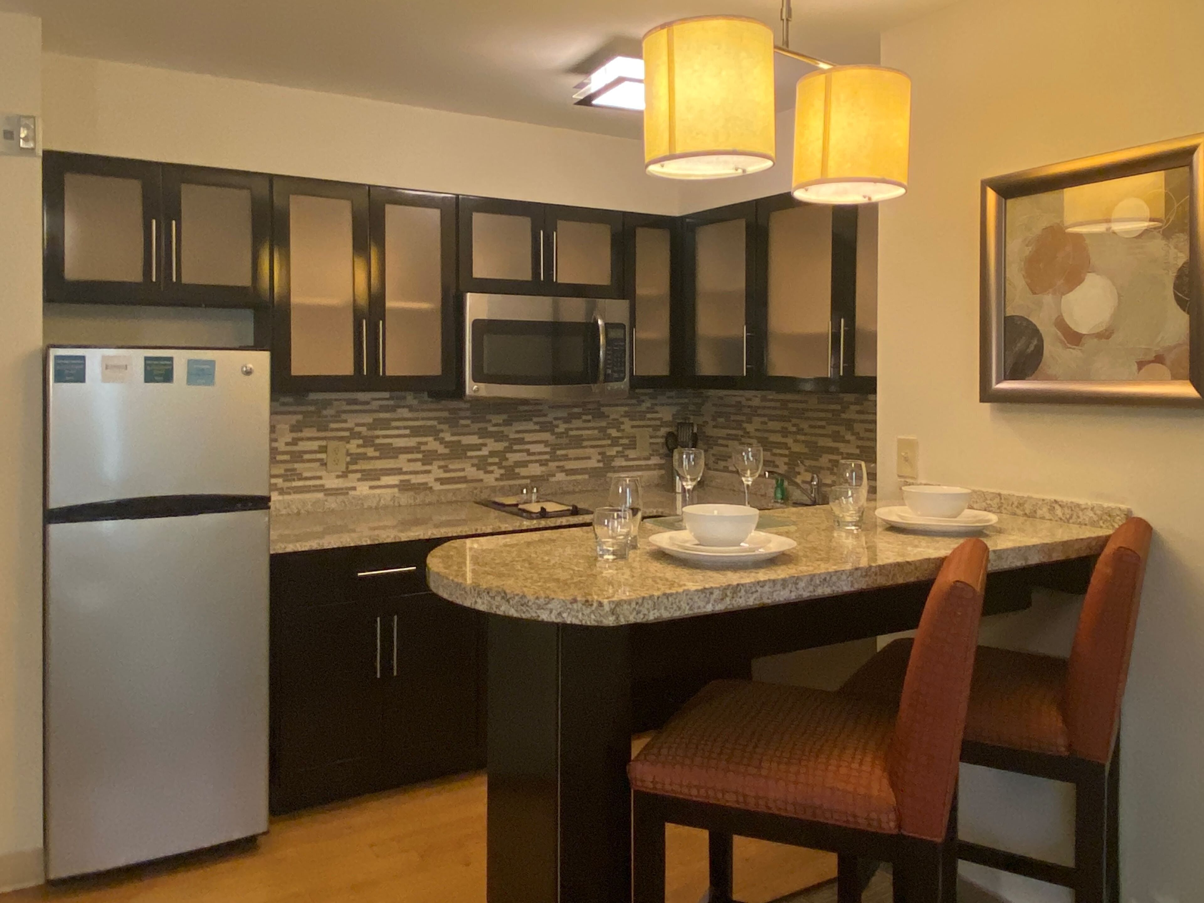Cocina compacta con armarios oscuros, microondas, refrigerador y comedor con mesa y sillas.