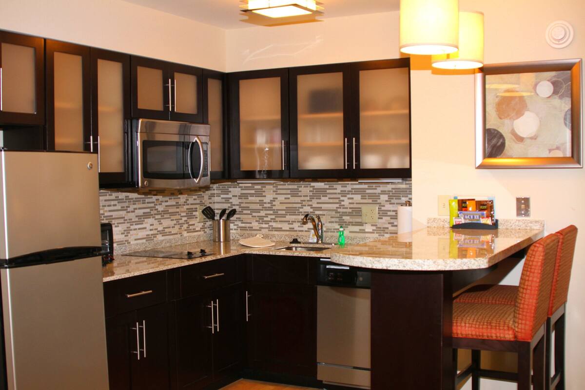 A modern kitchen with dark cabinets, stainless steel appliances, a granite countertop, and a bar area with a red cushioned chair.