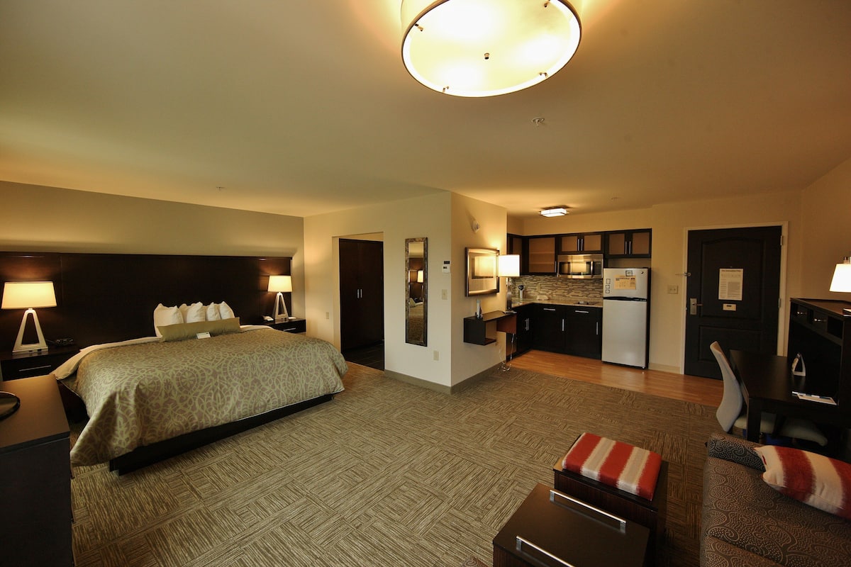 A hotel room with a bed, bedside tables, a desk, a chair, a kitchenette, and a bathroom.