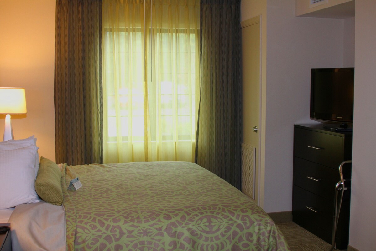 Une chambre d’hôtel avec un lit, un téléviseur posé sur une commode et une fenêtre avec des rideaux.