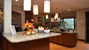 A hotel reception area with a granite counter, a flower arrangement, and a view of the lobby through a glass partition. - Staybridge Suites Liverpool Syracuse by IHG (Liverpool)