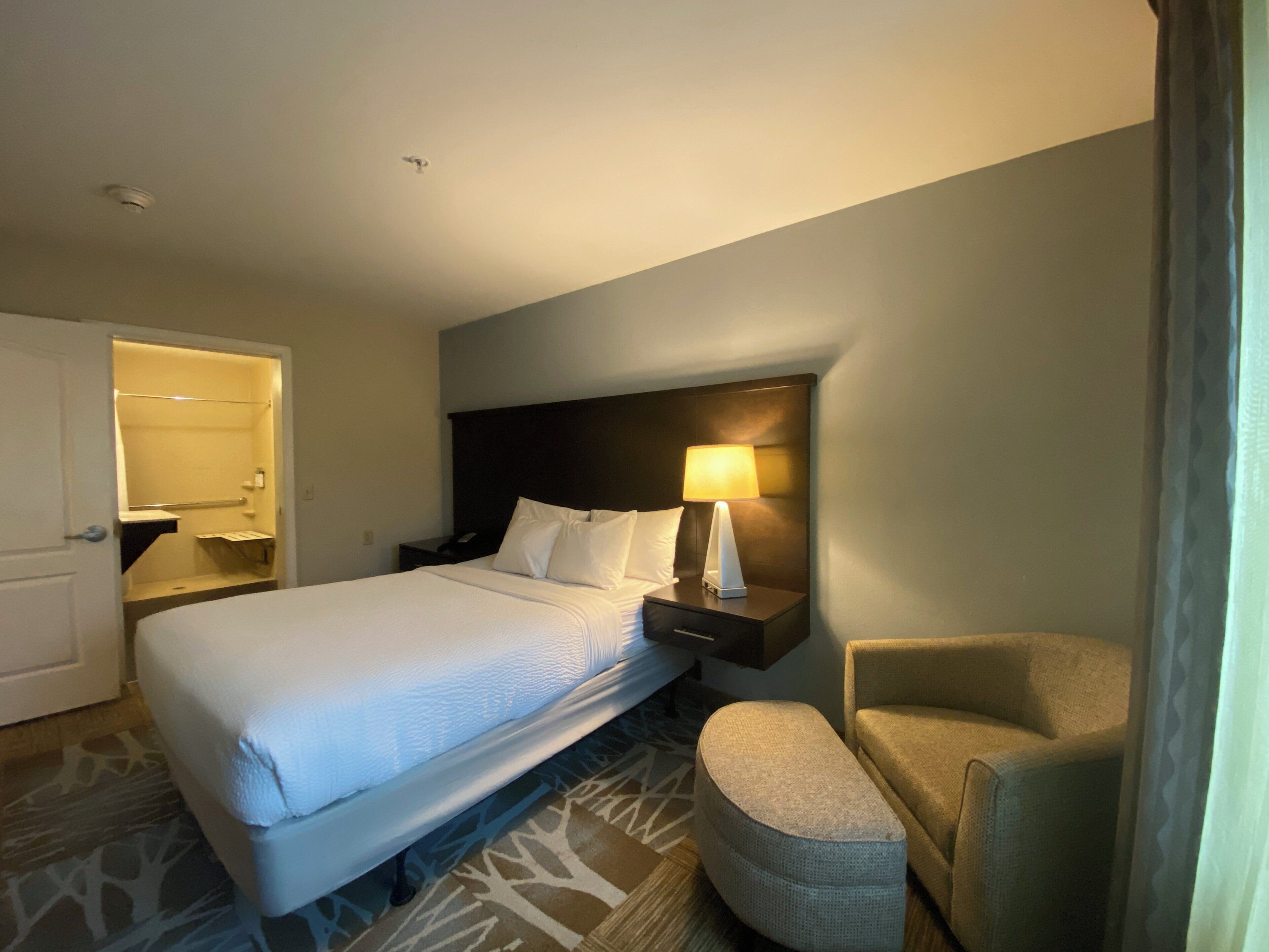 Habitación de hotel con cama, mesita de noche, lámpara, silla y puerta de baño.