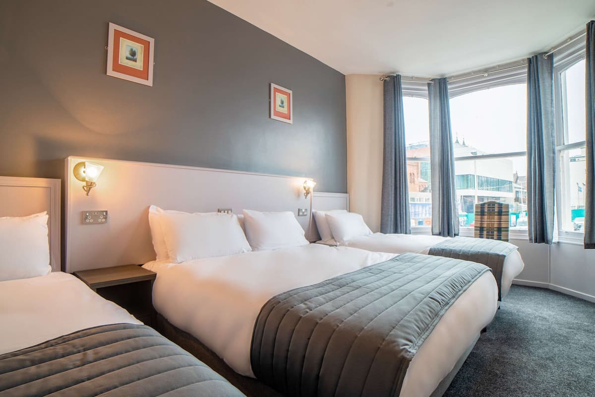 Une chambre d’hôtel avec trois lits, une grande fenêtre avec des rideaux et deux appliques murales.