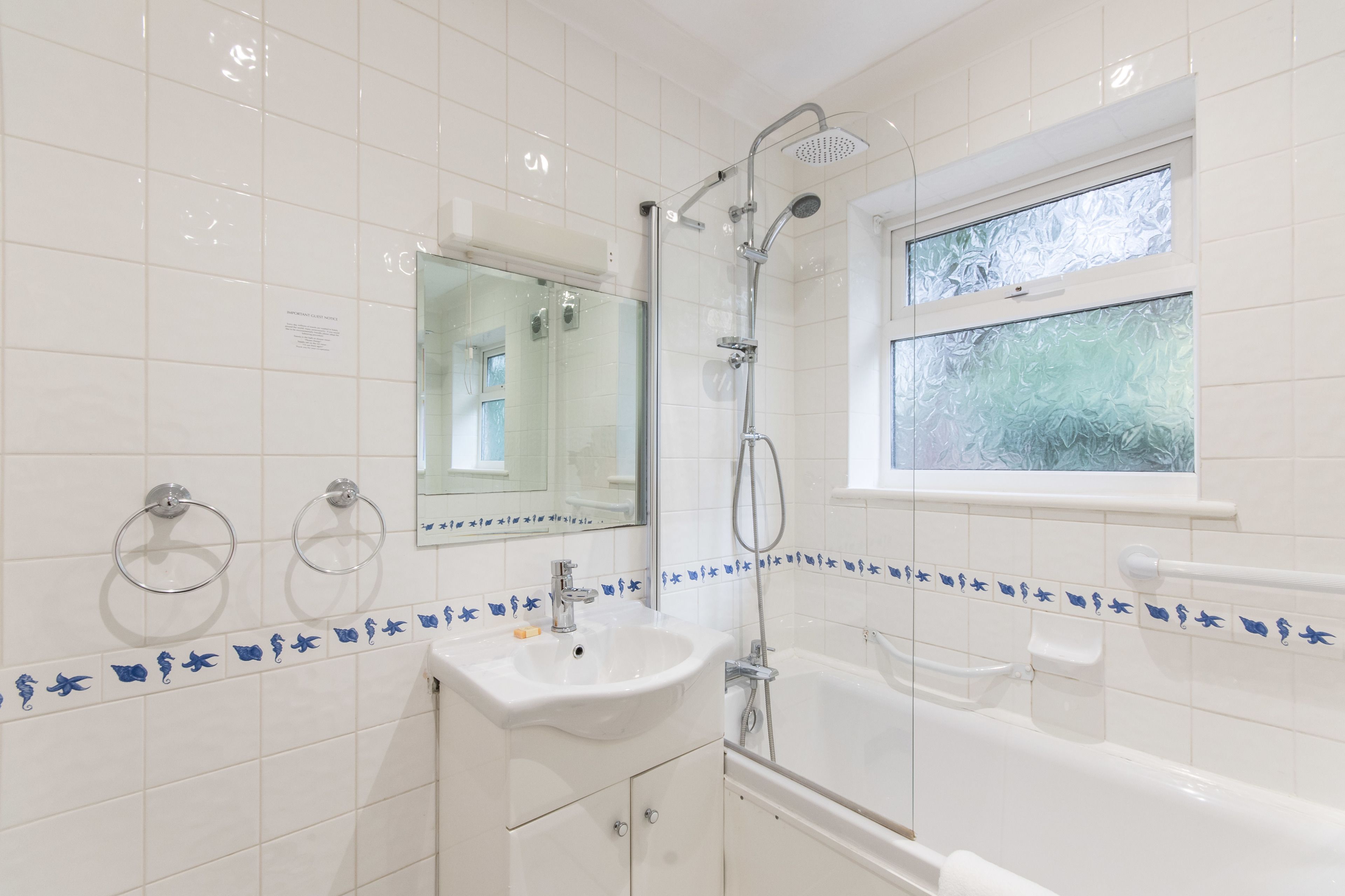 A bathroom with a white sink, mirror, and bathtub. There is a window with frosted glass and a showerhead.